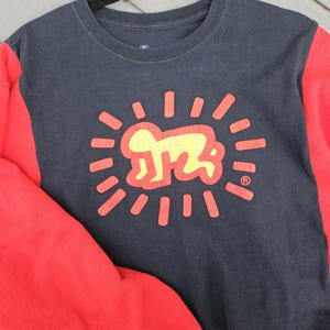Puede incluir: Una sudadera negra de cuello redondo con mangas rojas. La parte delantera presenta un gr&aacute;fico amarillo y rojo de una figura estilizada dentro de l&iacute;neas radiantes. El dise&ntilde;o recuerda la obra de arte de Keith Haring.