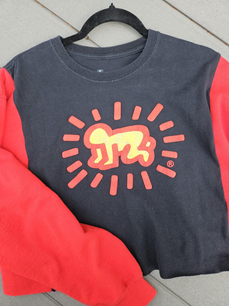 Puede incluir: Una sudadera negra de cuello redondo con mangas rojas. La parte delantera presenta un gr&aacute;fico amarillo y rojo de una figura estilizada dentro de l&iacute;neas radiantes. El dise&ntilde;o recuerda la obra de arte de Keith Haring.