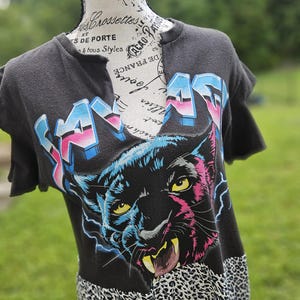 May include: A black short-sleeve t-shirt with a notched neckline featuring a graphic of a snarling black panther, the word SAVAGE in blue and pink gradient lettering, and a grey and black leopard print pattern along the bottom hem.