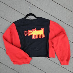 Puede incluir: Sudadera recortada negra y roja con un gr&aacute;fico amarillo de una figura similar a un perro. Las mangas son rojas y el cuerpo es negro con el gr&aacute;fico en el centro. La sudadera est&aacute; colgada de una percha negra.