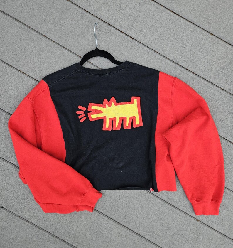 Puede incluir: Sudadera recortada negra y roja con un gr&aacute;fico amarillo de una figura similar a un perro. Las mangas son rojas y el cuerpo es negro con el gr&aacute;fico en el centro. La sudadera est&aacute; colgada de una percha negra.