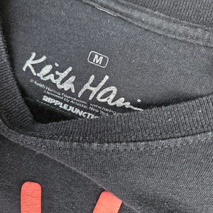 Puede incluir: Primer plano de una camiseta negra con el logotipo de la Fundaci&oacute;n Keith Haring y el texto "Keith Han" en blanco. La camiseta tiene una etiqueta de talla mediana. La parte inferior de la camiseta tiene un dise&ntilde;o rojo.