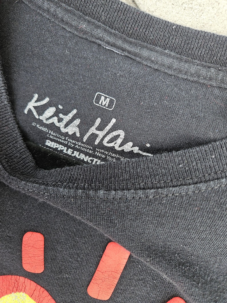 Puede incluir: Primer plano de una camiseta negra con el logotipo de la Fundaci&oacute;n Keith Haring y el texto "Keith Han" en blanco. La camiseta tiene una etiqueta de talla mediana. La parte inferior de la camiseta tiene un dise&ntilde;o rojo.