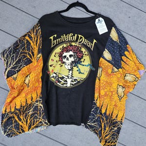 May include: A black t-shirt featuring a Grateful Dead graphic of a skull adorned with red roses inside a yellow circle. The sleeves are made of a contrasting fabric with a yellow and black pattern depicting bare trees and floral motifs.