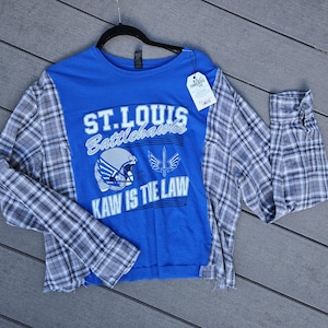 Pu&ograve; includere: Una maglia a maniche lunghe blu con una grafica dei St. Louis Battlehawks che include un casco da football, il logo di una spada e la scritta ST. LOUIS BATTLEHAWKS KAW IS THE LAW. La maglia presenta maniche in flanella a quadri grigi e bianchi.
