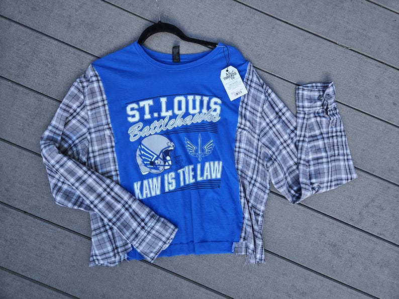 Pu&ograve; includere: Una maglia a maniche lunghe blu con una grafica dei St. Louis Battlehawks che include un casco da football, il logo di una spada e la scritta ST. LOUIS BATTLEHAWKS KAW IS THE LAW. La maglia presenta maniche in flanella a quadri grigi e bianchi.