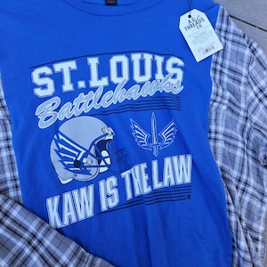 Pu&ograve; includere: Una t-shirt blu con il testo ST. LOUIS BATTLEHAWKS e KAW IS THE LAW, una grafica di un casco da football e un emblema a forma di spada. La maglia presenta maniche a quadri grigi e bianchi e un'etichetta A.G. THREADS CO. attaccata al colletto.