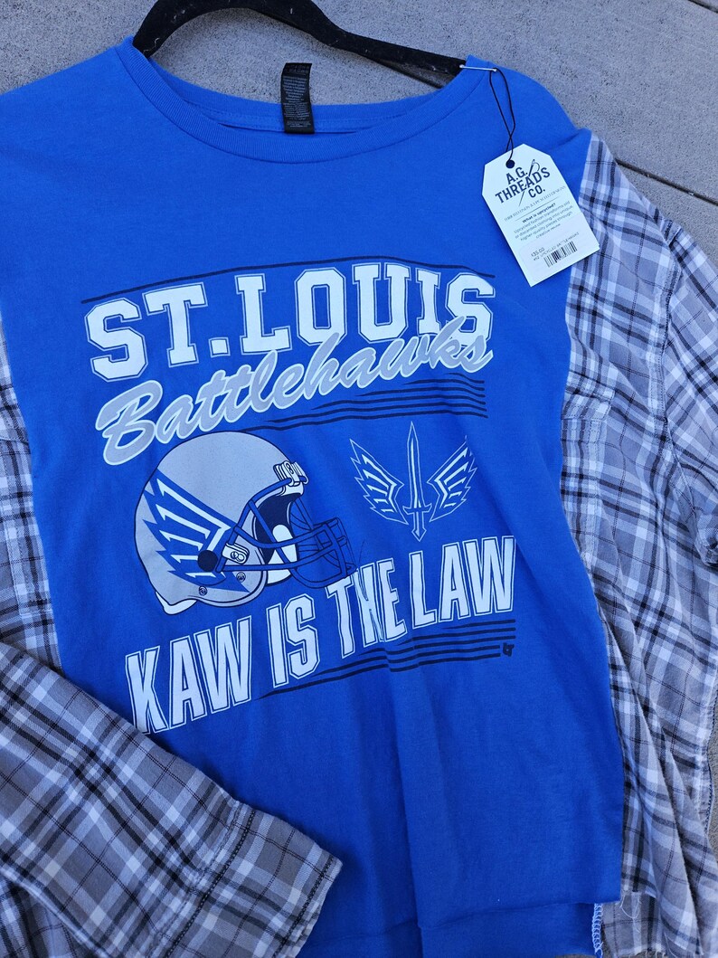 Pu&ograve; includere: Una t-shirt blu con il testo ST. LOUIS BATTLEHAWKS e KAW IS THE LAW, una grafica di un casco da football e un emblema a forma di spada. La maglia presenta maniche a quadri grigi e bianchi e un'etichetta A.G. THREADS CO. attaccata al colletto.