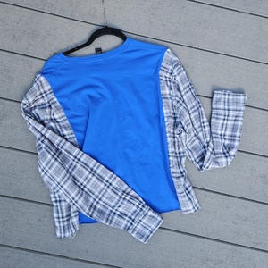 Pu&ograve; includere: Una maglia a maniche lunghe con un pannello centrale in jersey blu brillante, a contrasto con le maniche e i pannelli laterali caratterizzati da un motivo a quadri bianco, grigio e nero. Il capo &egrave; appeso a una gruccia in plastica nera.