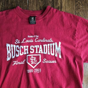 May include: A red t-shirt featuring the text "BUSCH STADIUM" in large white letters, with "St. Louis Cardinals" above and "Final Season 1966-2005" below. The shirt has a crew neck and a small tag.