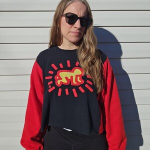 Puede incluir: Una sudadera corta negra y roja con una figura amarilla y l&iacute;neas rojas radiantes. La sudadera tiene mangas largas rojas y un torso negro con el gr&aacute;fico. La persona lleva gafas de sol negras y leggings negros.