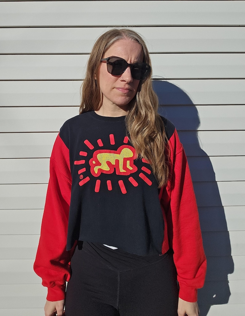 Puede incluir: Una sudadera corta negra y roja con una figura amarilla y l&iacute;neas rojas radiantes. La sudadera tiene mangas largas rojas y un torso negro con el gr&aacute;fico. La persona lleva gafas de sol negras y leggings negros.