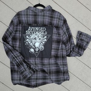 May include: A gray and black plaid flannel shirt with a large patch featuring the text "AVENGED SEVENFOLD" and a skull graphic. The shirt is hanging on a black hanger.