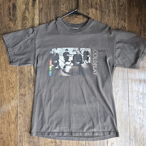 May include: A gray t-shirt featuring a band photo and the word "COLDPLAY" printed vertically on the right side. The band photo includes a colorful stripe on the left side. The shirt is hanging on a black hanger.