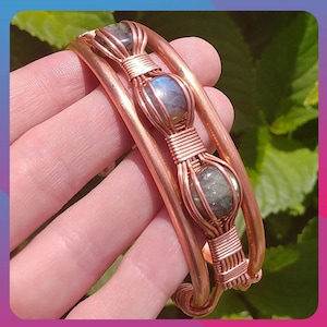 May include: A copper wire cuff bracelet with three labradorite stones wrapped in wire. The bracelet is shown on a hand.