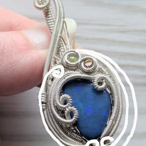 以下が含まれることがあります： 鮮やかなブルーオパールをあしらった手作りのペンダント。 オパールは、繊細な渦巻きとループを持つ複雑な銀線で囲まれています。 2つの小さな宝石がオパールの上にセットされ、このユニークなジュエリーにディテールを加えています。