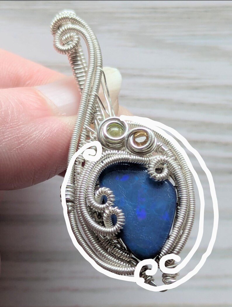 以下が含まれることがあります： 鮮やかなブルーオパールをあしらった手作りのペンダント。 オパールは、繊細な渦巻きとループを持つ複雑な銀線で囲まれています。 2つの小さな宝石がオパールの上にセットされ、このユニークなジュエリーにディテールを加えています。