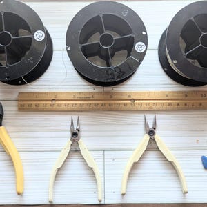 以下が含まれることがあります： ジュエリー作りのツールと材料の俯瞰図。黒いワイヤーの3つのスプール、インチでマークされた木製の定規、黄とクリーム色のハンドルの3組のペンチ、小さな青い石が含まれています。定規の長さは30.5cmです。