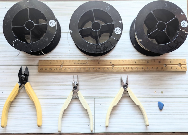 以下が含まれることがあります： ジュエリー作りのツールと材料の俯瞰図。黒いワイヤーの3つのスプール、インチでマークされた木製の定規、黄とクリーム色のハンドルの3組のペンチ、小さな青い石が含まれています。定規の長さは30.5cmです。