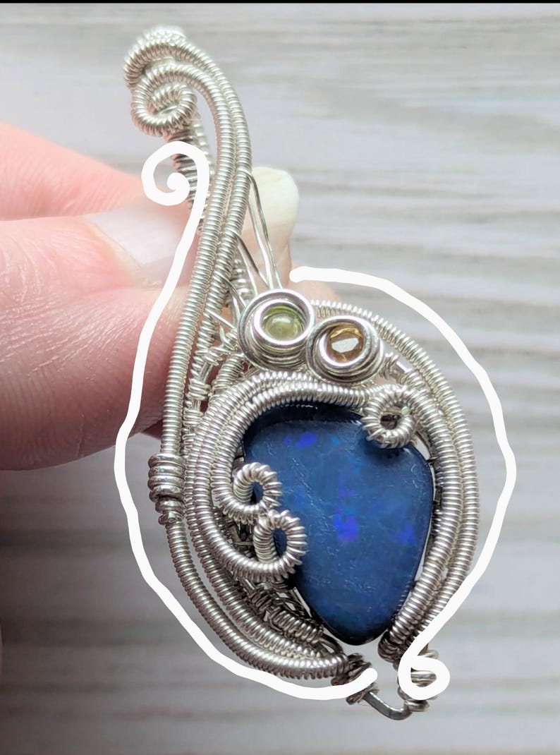 以下が含まれることがあります： 鮮やかな青色のオパール石を銀線で精巧に包んだ手作りのペンダント。銀線は石の周りに装飾的なフレームを形成し、小さな緑色と茶色の石がアクセントになっています。このペンダントはユニークで芸術的なデザインです。
