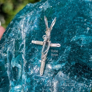 May include: A silver wire-wrapped cross pendant with a clear stone center. The cross is displayed on a textured, teal-colored surface. The text "Tobe Rose" is visible on the surface.