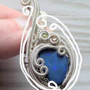 以下が含まれることがあります： 鮮やかな青色のオパール宝石をあしらった、手作りの銀線巻きペンダント。ペンダントは、渦巻き模様と小さな緑と茶色のアクセントストーンを備えた複雑なワイヤーワークが特徴です。オパールは色の遊びを示し、そのユニークな魅力を高めています。