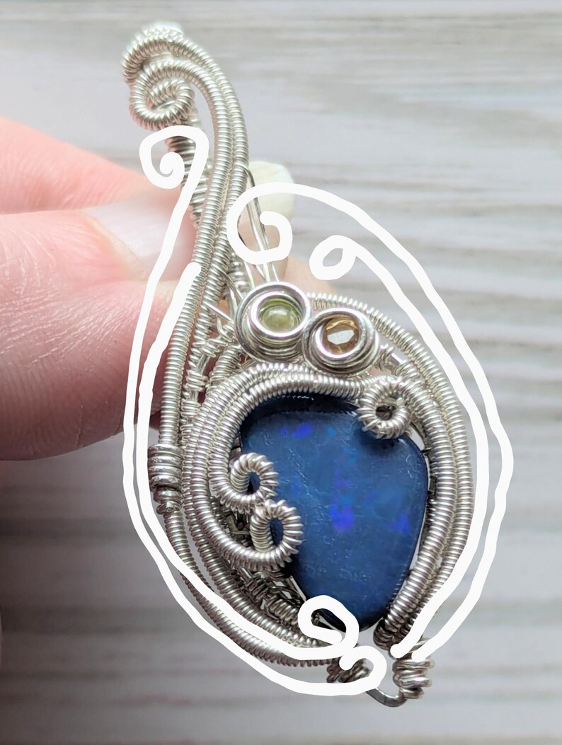 以下が含まれることがあります： 鮮やかな青色のオパール宝石をあしらった、手作りの銀線巻きペンダント。ペンダントは、渦巻き模様と小さな緑と茶色のアクセントストーンを備えた複雑なワイヤーワークが特徴です。オパールは色の遊びを示し、そのユニークな魅力を高めています。