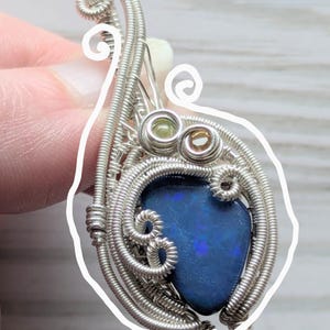 以下が含まれることがあります： 鮮やかなブルーオパールストーンをあしらった、手作りのシルバーワイヤーラップペンダント。ペンダントは、小さな緑と茶色のアクセントストーンを備えた、複雑なワイヤーディテールが特徴です。ユニークで芸術的なジュエリーです。