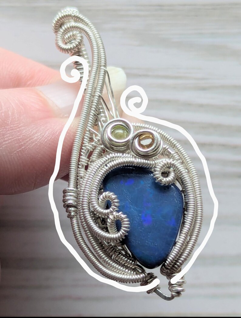 以下が含まれることがあります： 鮮やかなブルーオパールストーンをあしらった、手作りのシルバーワイヤーラップペンダント。ペンダントは、小さな緑と茶色のアクセントストーンを備えた、複雑なワイヤーディテールが特徴です。ユニークで芸術的なジュエリーです。
