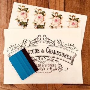 Puede incluir: Una hoja de papel blanca con un diseño gráfico en blanco y negro de un letrero vintage que dice "Manufacture de Chaussures" con el texto "Vissées et Gondées en 1835" debajo. Una herramienta raspadora de plástico azul está encima del papel. El papel está encima de una hoja de papel con cuatro diseños florales.