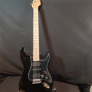 Puede incluir: Guitarra eléctrica negra con diapasón de arce y herrajes cromados. La guitarra está en un soporte.