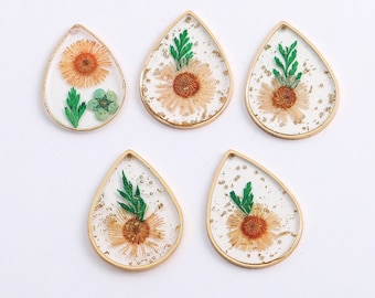 May include: Five teardrop-shaped pendants with gold-colored frames. Each pendant contains a dried flower and greenery encased in clear resin. The flowers are primarily orange and yellow, with green accents. The pendants are displayed on a white surface.