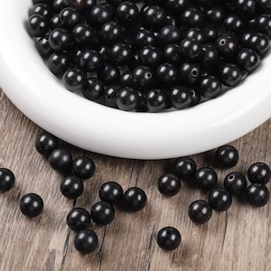 May include: A collection of black, round beads, some scattered on a wooden surface, and the rest in a white, shallow bowl. The beads have a glossy finish and are evenly sized, suggesting they are for jewellery making or crafts.