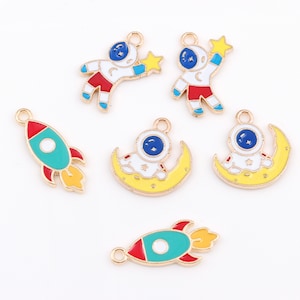 Puede incluir: Seis abalorios de metal dorado con pintura de esmalte. Los abalorios presentan diseños de astronautas y cohetes en rojo, azul, amarillo, blanco y verde.