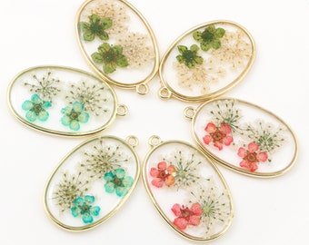 May include: Six oval-shaped gold-rimmed pendants with pressed flowers and leaves inside. The pendants have a loop at the top for hanging. The flowers are in shades of green, pink, and blue.