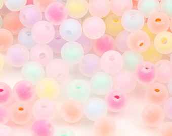 Puede incluir: Primer plano de una colección de cuentas de vidrio de color pastel. Las cuentas son redondas y tienen un acabado mate. Están esparcidas sobre una superficie blanca.