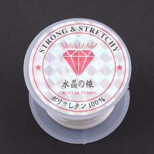 Puede incluir: Un carrete de hilo el&aacute;stico blanco etiquetado como "STRONG & STRETCHY" con un logotipo de diamante rojo y el texto "水晶の線 CRYSTAL STRING ポリウレタン 100%"