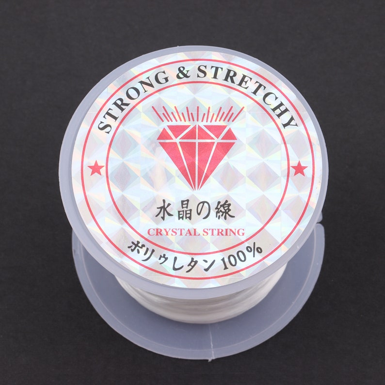 Puede incluir: Un carrete de hilo el&aacute;stico blanco etiquetado como "STRONG & STRETCHY" con un logotipo de diamante rojo y el texto "水晶の線 CRYSTAL STRING ポリウレタン 100%"