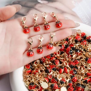 Peut inclure: Gros plan sur des breloques coccinelles avec des détails en émail rouge et noir, des apprêts en métal doré et des fermoirs mousquetons. Les breloques sont présentées avec une pile de breloques similaires, suggérant un thème de fabrication de bijoux.