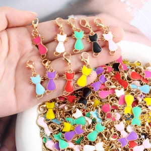 May include: A collection of gold-tone metal cat-shaped charms with colourful enamel fill, each featuring a lobster claw clasp. The charms come in various colours including pink, white, green, black, light blue, purple, red, and yellow.