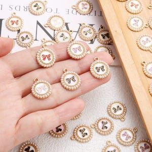 May include: Gold-coloured round charms with a pearl border and a bow design in various colours. The charms have a small loop at the top for attaching to jewellery or other crafts. The bows are in red, black, pink, and white.