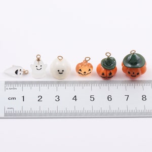 Może przedstawiać: Sześć uroczych charm&oacute;w na Halloween, w tym duchy i dynie, ze złotymi k&oacute;łkami do zawieszenia. Charms są ułożone na linijce, kt&oacute;ra mierzy około 3,8 cm długości.
