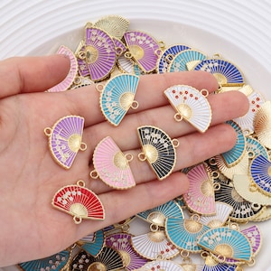 Może przedstawiać: Zestaw kolorowych metalowych zawieszek w kształcie wachlarza z złotymi akcentami i białymi detalami emaliowanymi. Zawieszki mają pętelkę na górze do przymocowania do biżuterii lub innych prac rękodzielniczych.