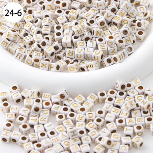May include: White plastic letter beads with gold lettering. The beads are in a white bowl and scattered on a white surface. The beads spell out the letters of the alphabet.