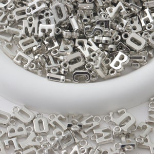 May include: A large pile of silver metal letter charms in a white bowl. The charms are all different letters of the alphabet and are perfect for jewelry making or crafting.