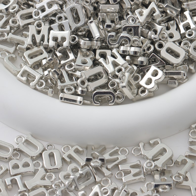 May include: A large pile of silver metal letter charms in a white bowl. The charms are all different letters of the alphabet and are perfect for jewelry making or crafting.