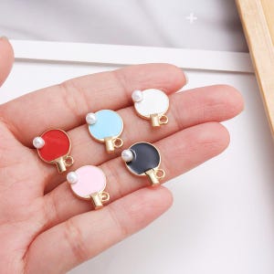 Puede incluir: Cinco colgantes de raquetas de ping pong de colores. Cada colgante presenta una raqueta de diferente color: rojo, azul claro, blanco, negro y rosa. Una pequeña perla blanca está unida a cada raqueta. Las raquetas tienen mangos dorados.