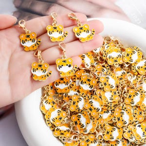 Puede incluir: Encantos de tigre dorados con detalles de esmalte naranja y negro. Los amuletos tienen la forma de un cachorro de tigre con un cierre dorado. Un cuenco blanco está lleno de amuletos similares.