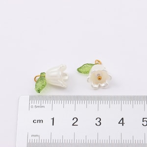 Puede incluir: Dos abalorios de flores blancas con hojas verdes y anillas de salto doradas. Los abalorios miden aproximadamente 1 cm y se muestran junto a una regla.