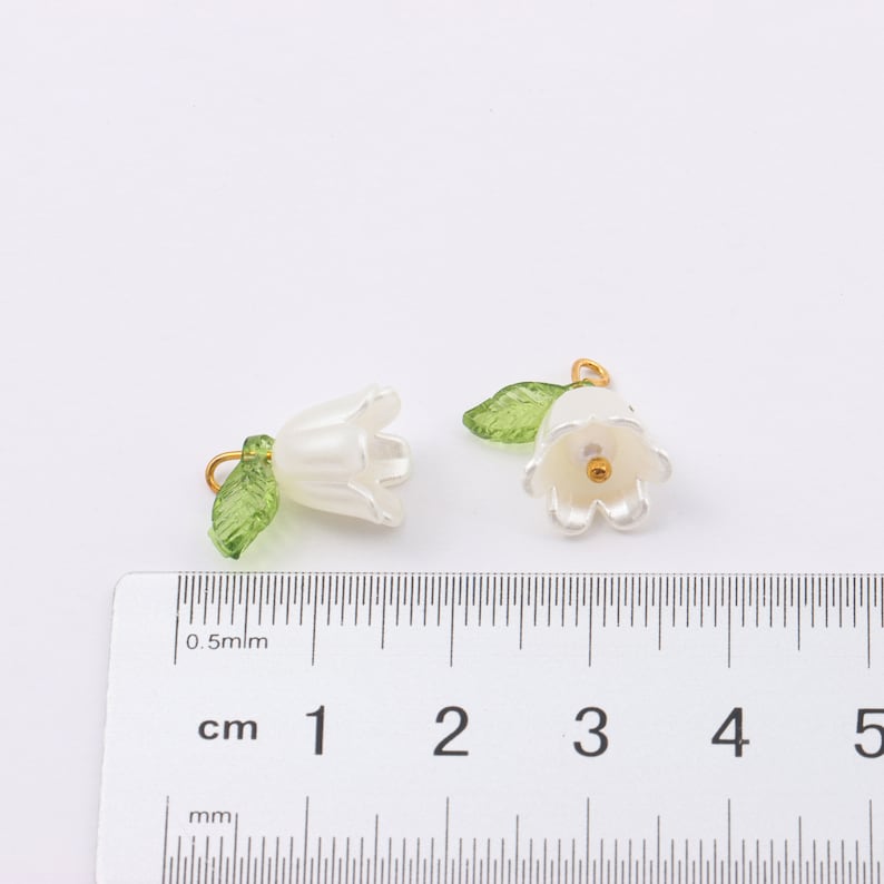 Puede incluir: Dos abalorios de flores blancas con hojas verdes y anillas de salto doradas. Los abalorios miden aproximadamente 1 cm y se muestran junto a una regla.